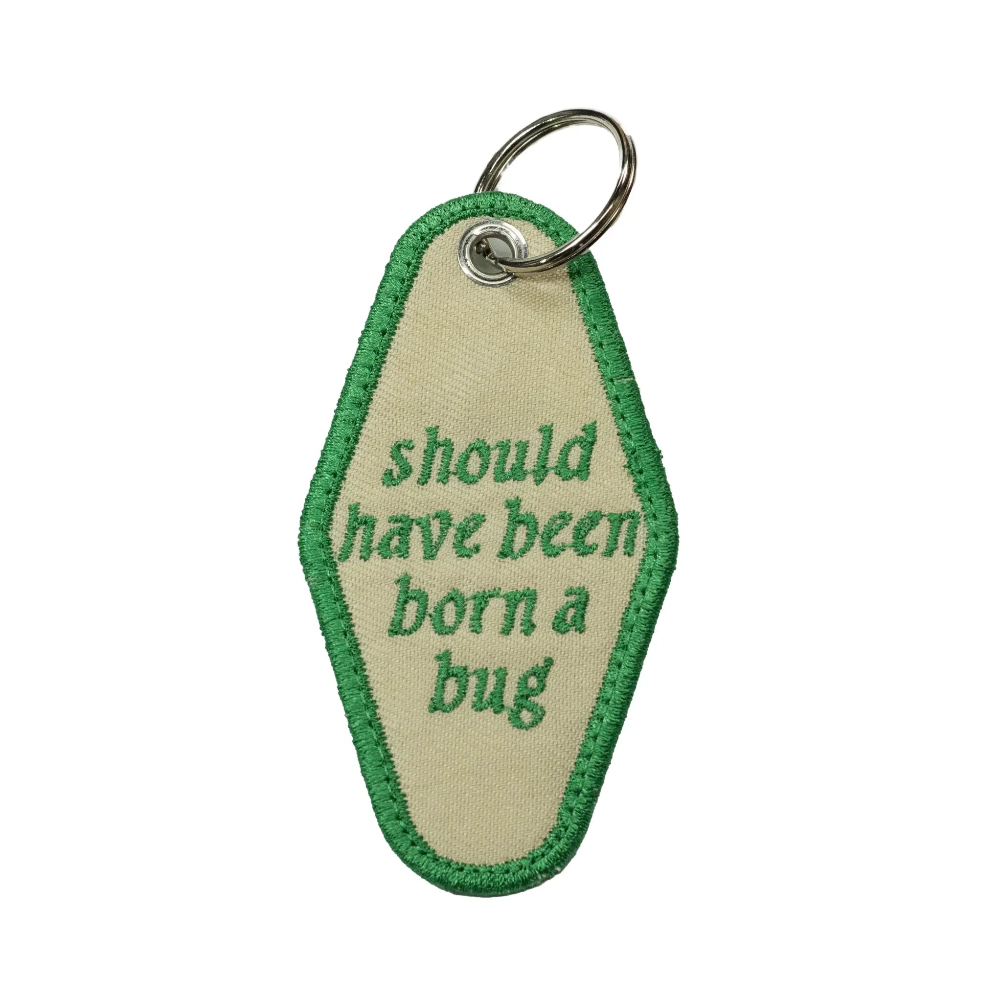 Back view of a beige diamond-shaped motel keychain by Regular Magic featuring green embroidered text that reads 'should have been born a bug' with a green merrowed border.