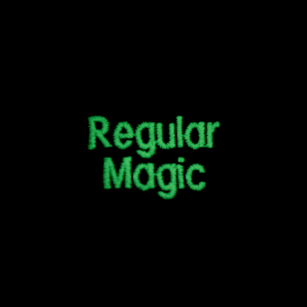 Close-up of the Regular Magic logo glowing in the dark with bright green embroidered thread against a black background.