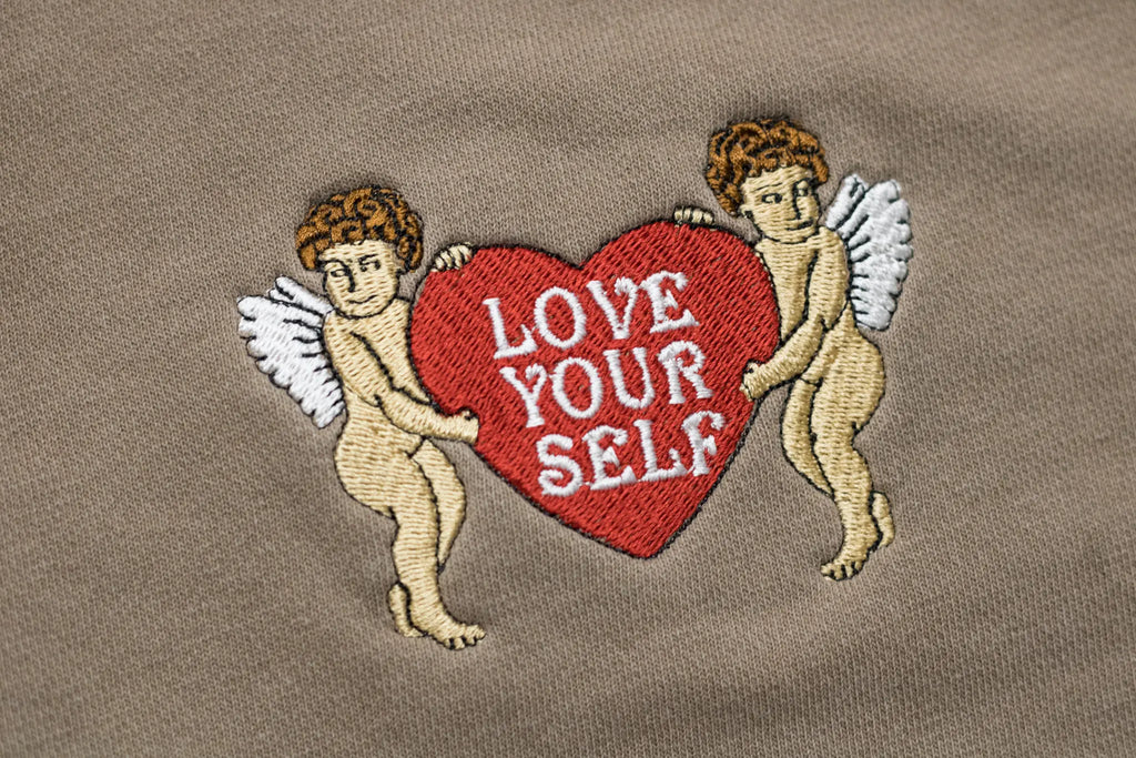Close-up detail of embroidery on a clay-colored garment featuring two cherubs holding a red heart with white text reading 'LOVE YOUR SELF'.