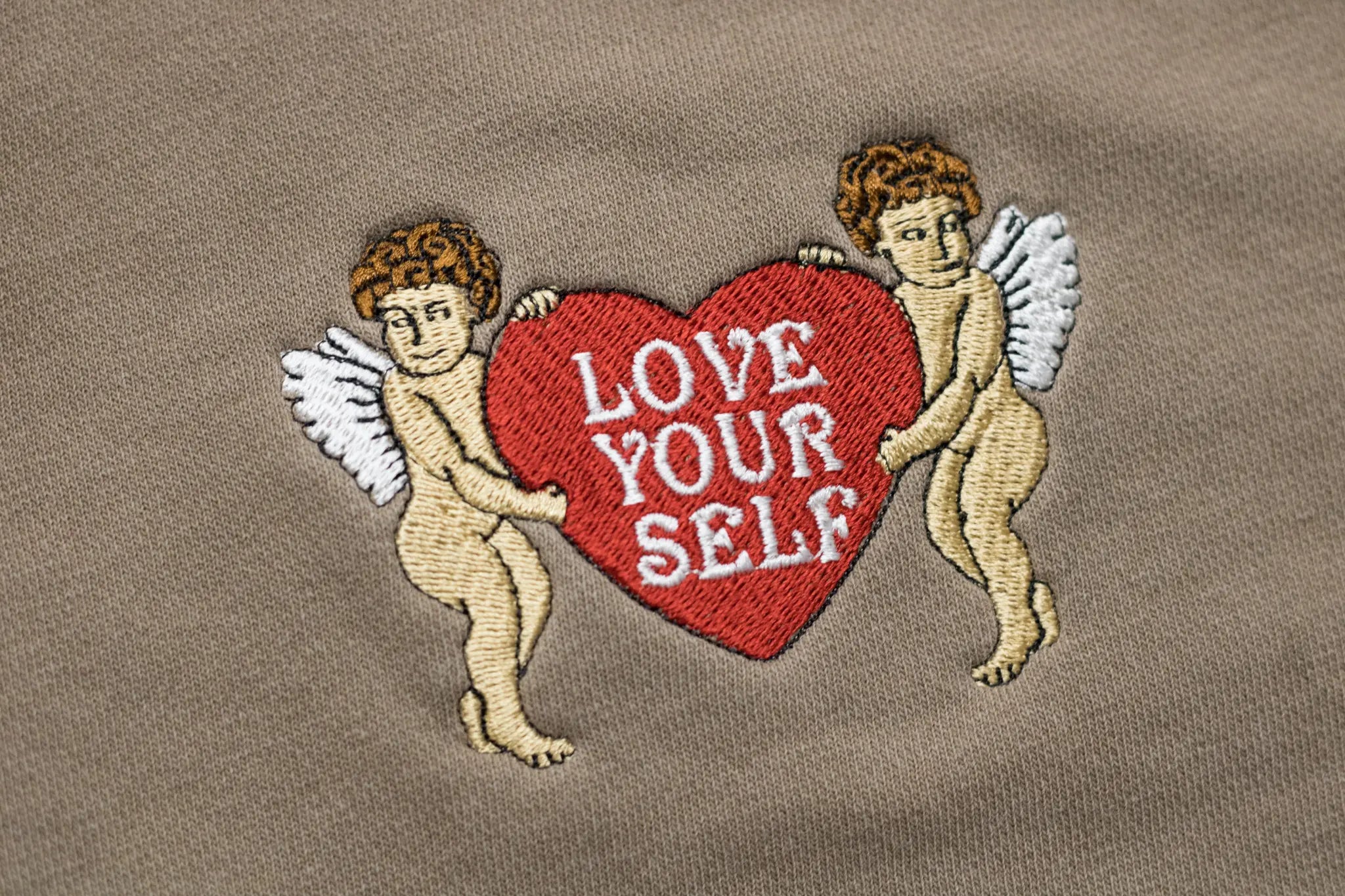 Close-up detail of embroidery on a clay-colored garment featuring two cherubs holding a red heart with white text reading 'LOVE YOUR SELF'.
