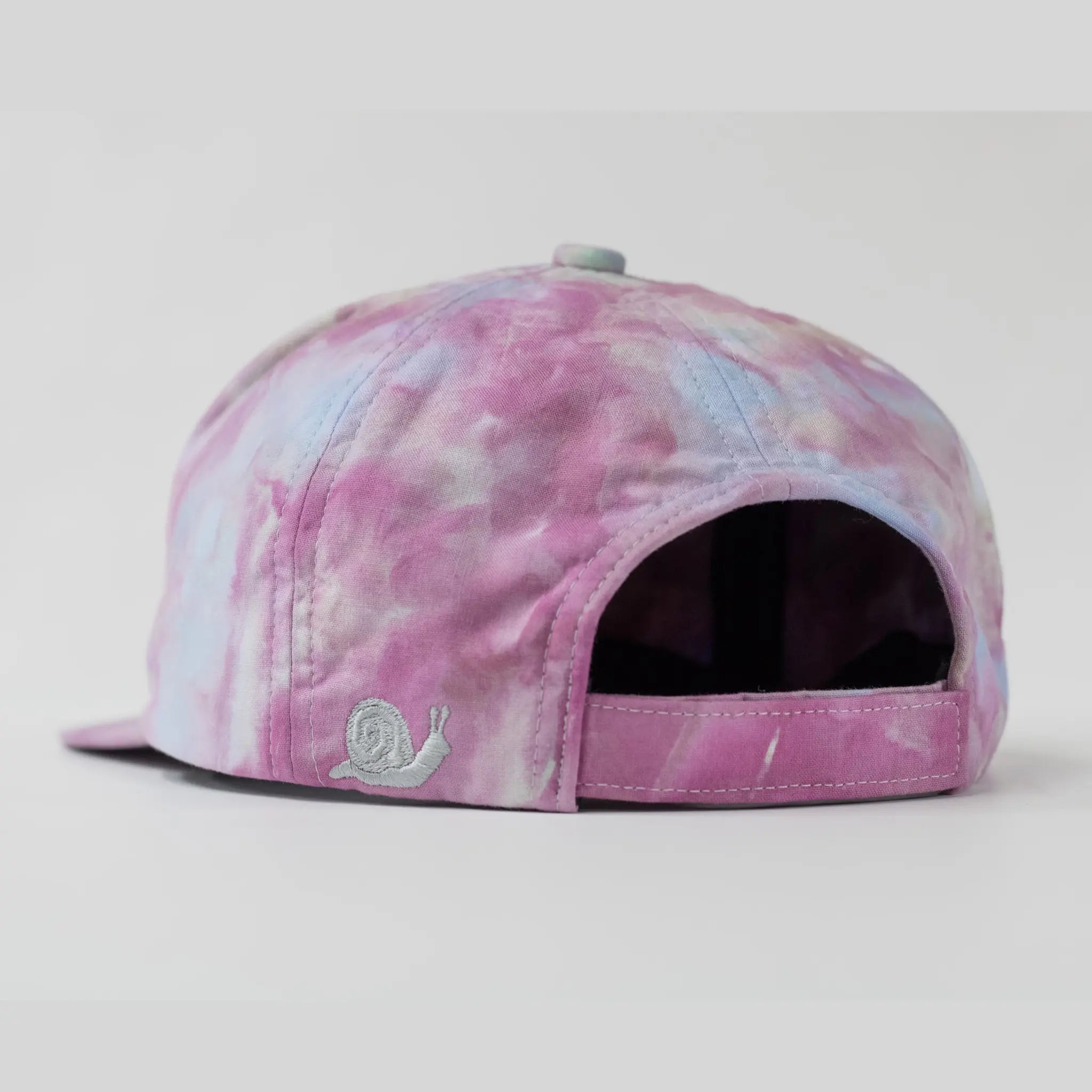 Back view of a Regular Magic pink and blue pastel 6 panel baseball tie-dye cap showing an adjustable strap and a silver embroidered snail logo near the back edge.