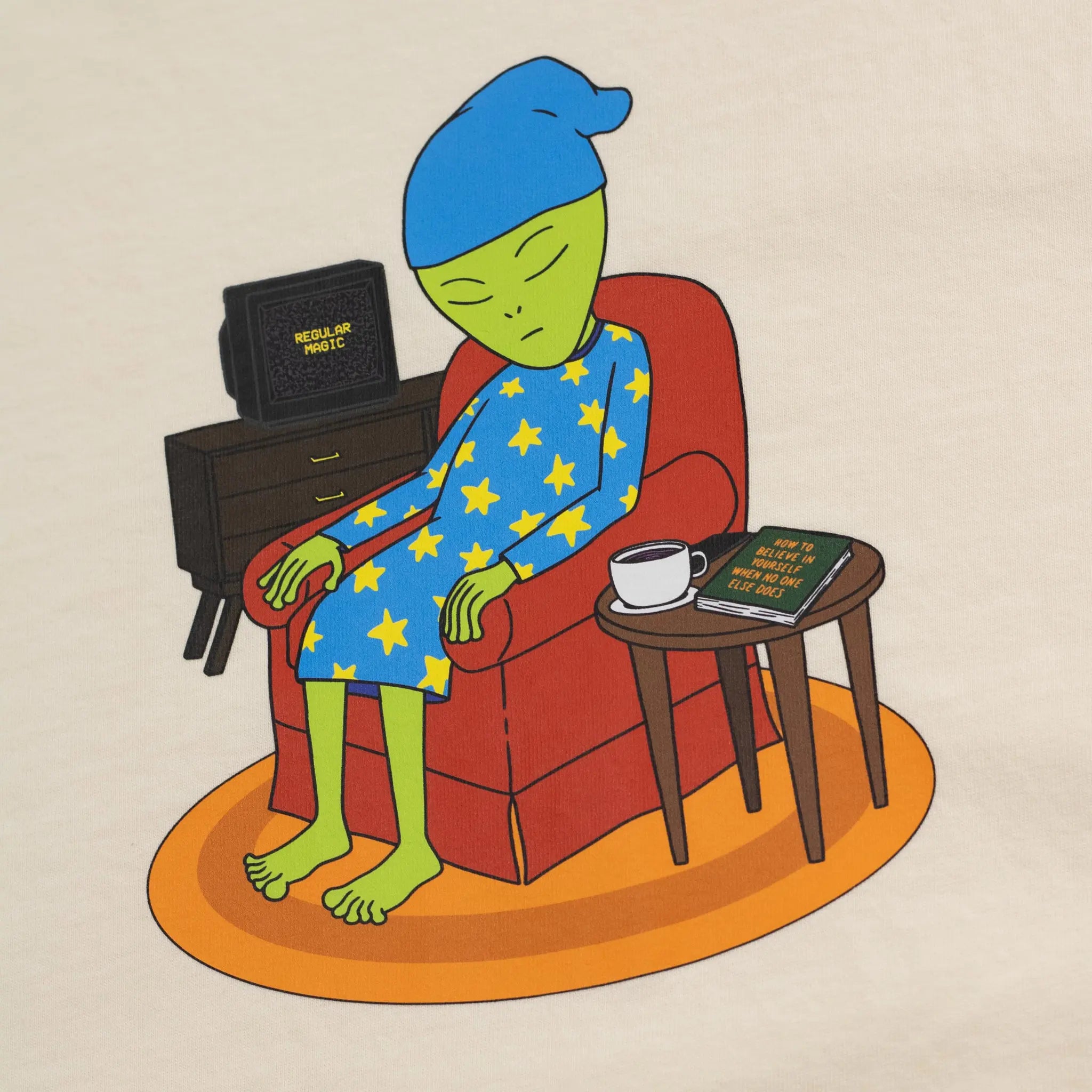 Close-up graphic on a beige Regular Magic t-shirt featuring the Sleepy Alien character sleeping in a red armchair, wearing star-patterned pajamas alongside a mug, TV, and a book titled 'How To Believe In Yourself When No One Else Does'.