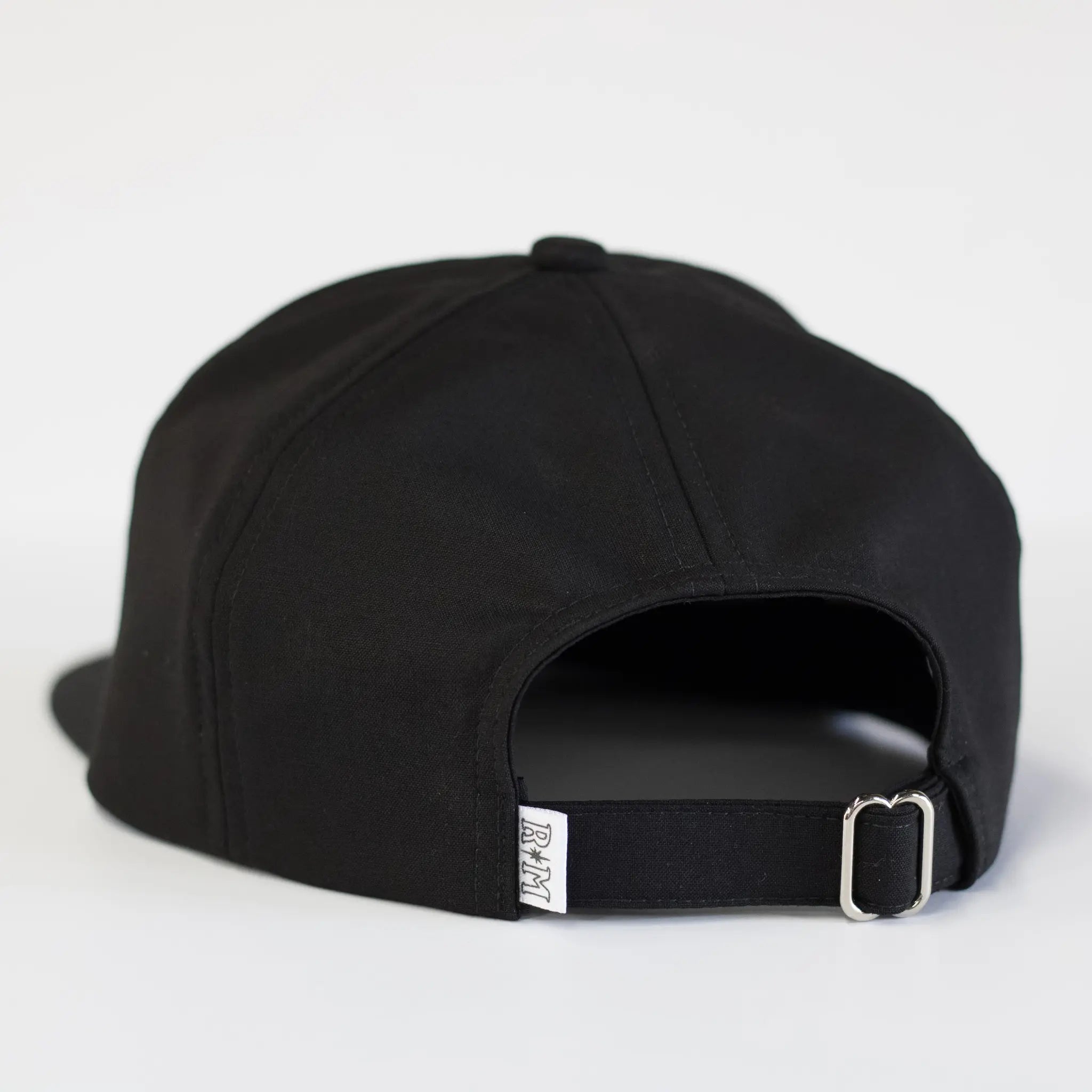 Back view of a black Regular Magic cap showing the adjustable fabric strap, metal buckle closure, and printed RM logo tag on the back brim.