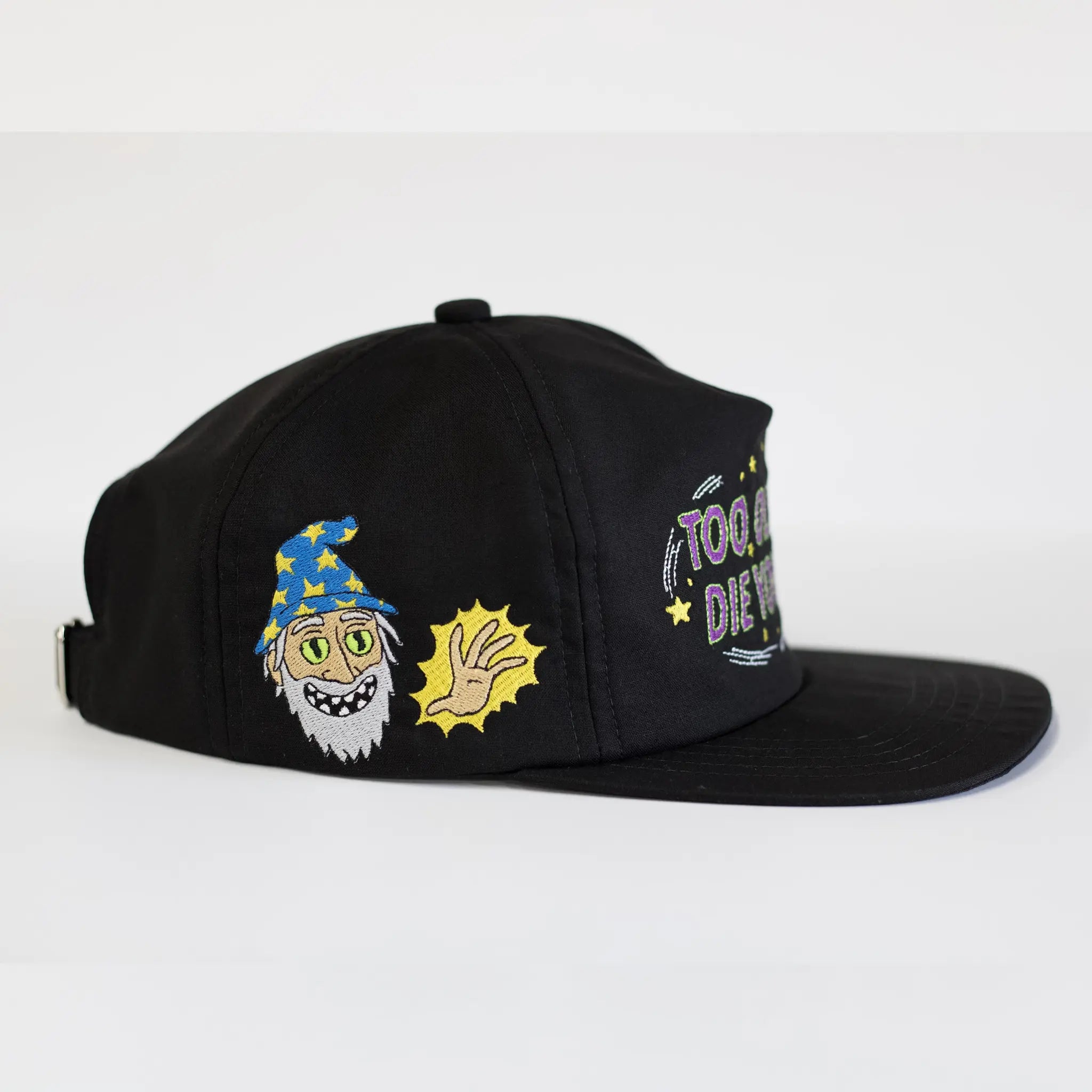 Side view of a black Regular Magic cap featuring embroidered front text reading 'TOO OLD TO DIE YOUNG' and a side patch of the Wizard character with a star hat next to a glowing magic hand.